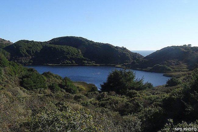 A small blue lake surrounded by brushy green hills with the ocean visible in the distance through a gap in the hills.