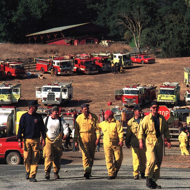 A crew of wildland firefighters wearing yellow clothing stride across a parking lot with a dozen fire trucks, a barn, and a forested ridge in the background.