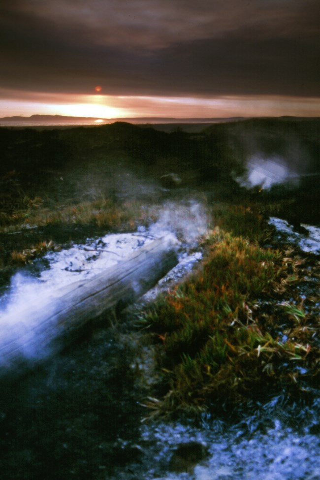 A smoking log and partially burnt grass in the foreground with the sun setting between low clouds and the horizon in the distance.