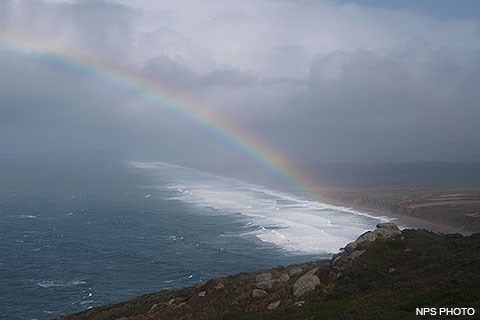 A rainbow arcs to the left above a stormy sea as big waves crash along a long straight beach on the right.