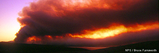 Smoke rises from a ridge on the left and drifts to the right as light from the sunrise in the distance filters through the smoke.