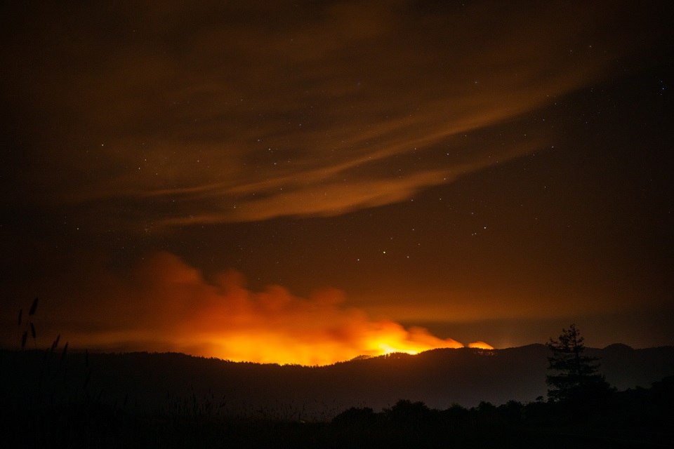 Smoke rises above a ridge and reflects orange light generated by a wildfire burning on the ridge's far side. Stars are visible above the smoke.