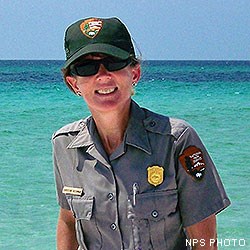 A head photo of Point Reyes Social Media Team member Christine wearing a gray ranger shirt and a ranger ballcap with turquoise-colored waters in the background.