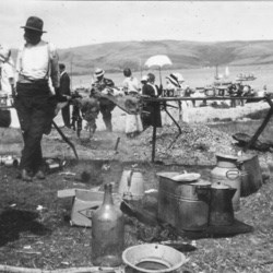 Black and white photo of people picnicking on a beach with water and hills in the background.
