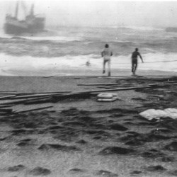 Black and white photo of two men on a beach with a ship surrounded by heavy surf very close to shore.