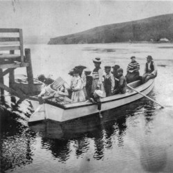 Black and white photo of people on a rowboat launching from a dock on the left. Low headlands rise above the far end of the bay.