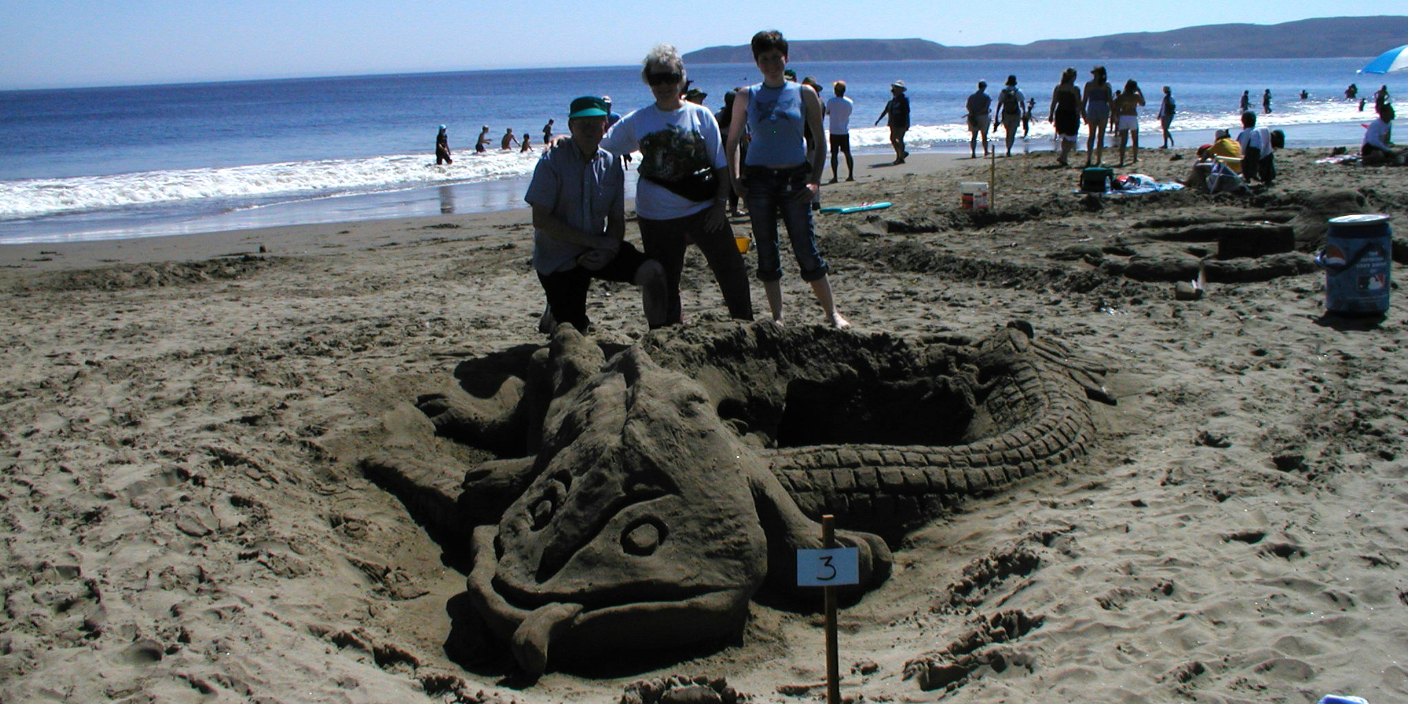 Three adults pose behind a sand sculpture of a salamander.