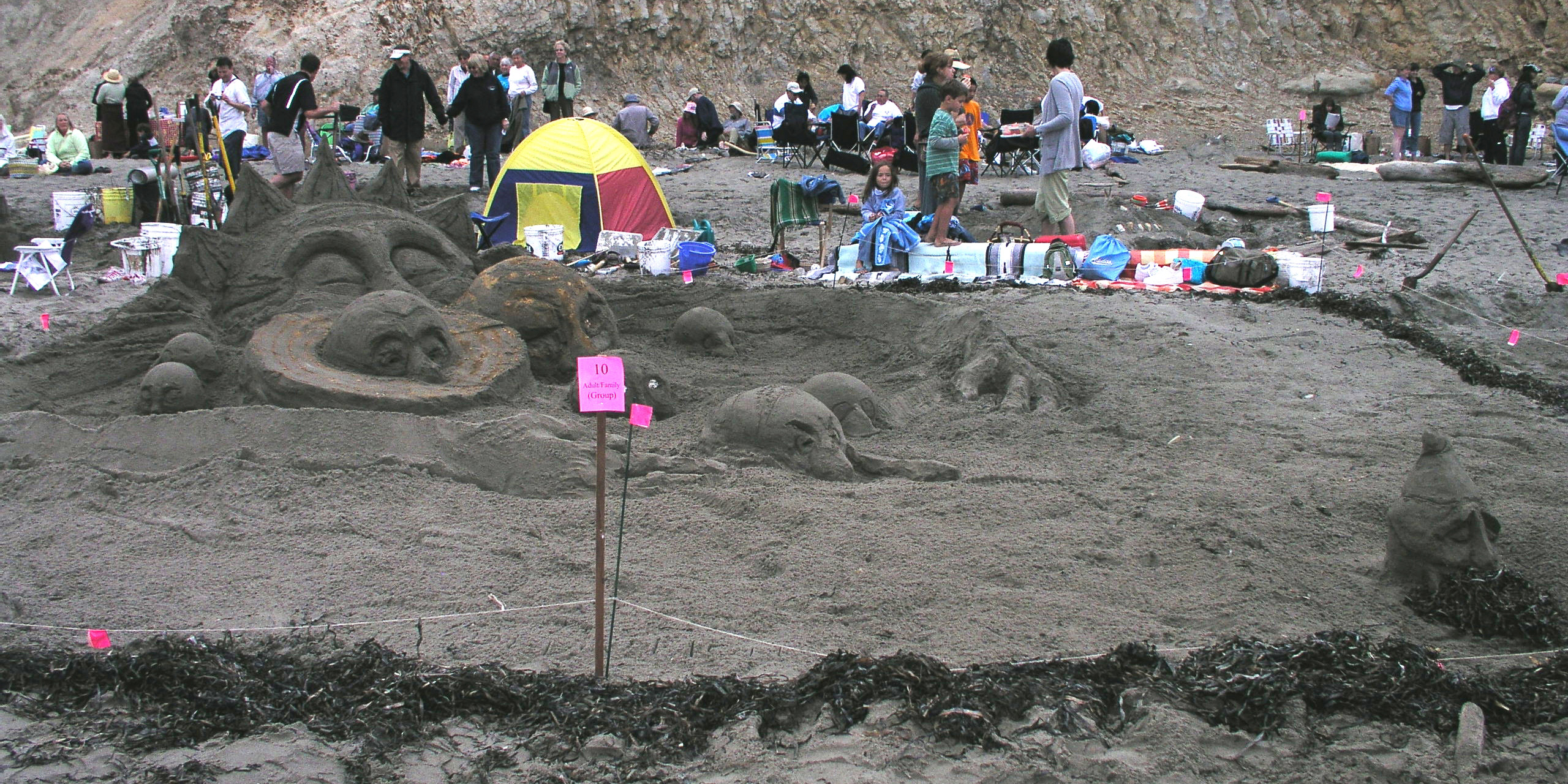 A large extensive sand sculpture of an anthropomorphic sun with its arms embracing eight anthropomorphic planets with a dwarvish-looking planet outside the sun's arms.