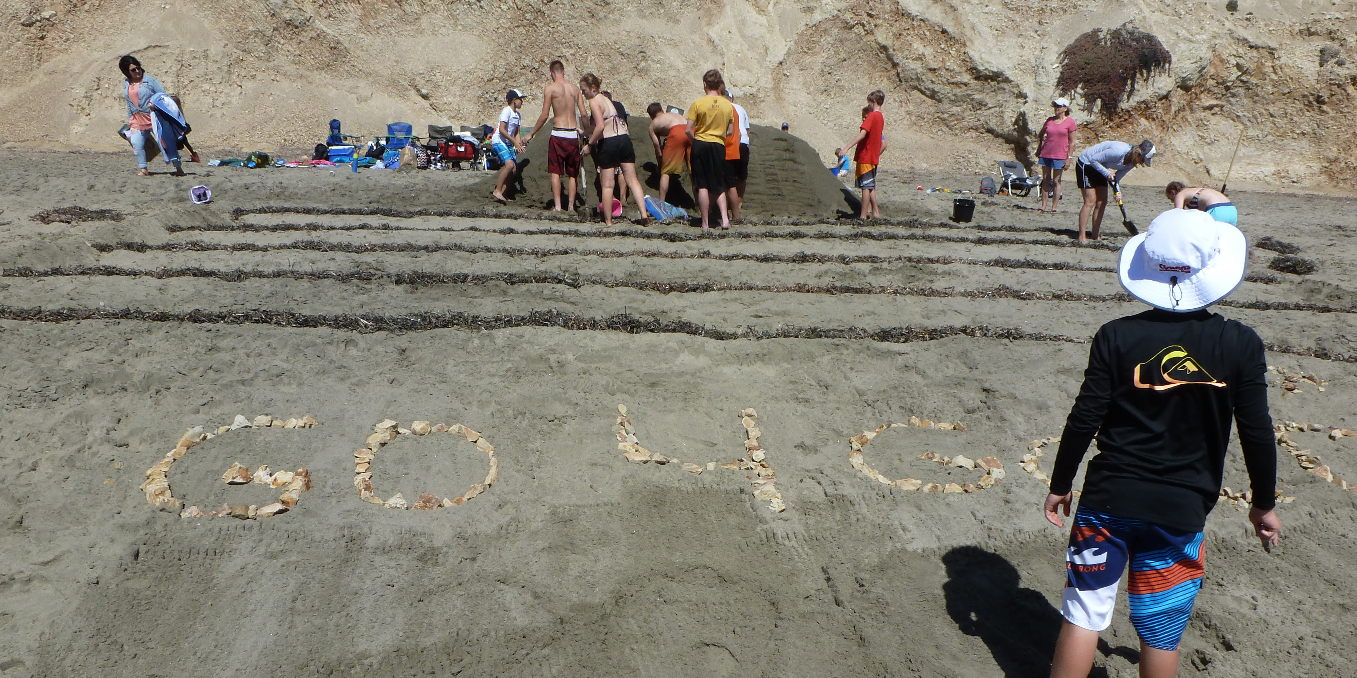 Jen Jordan and ten friends constructing bleachers of sand beyond a race track made of kelp. In the foreground, tan-colored rocks spell out Go 4 Gold.