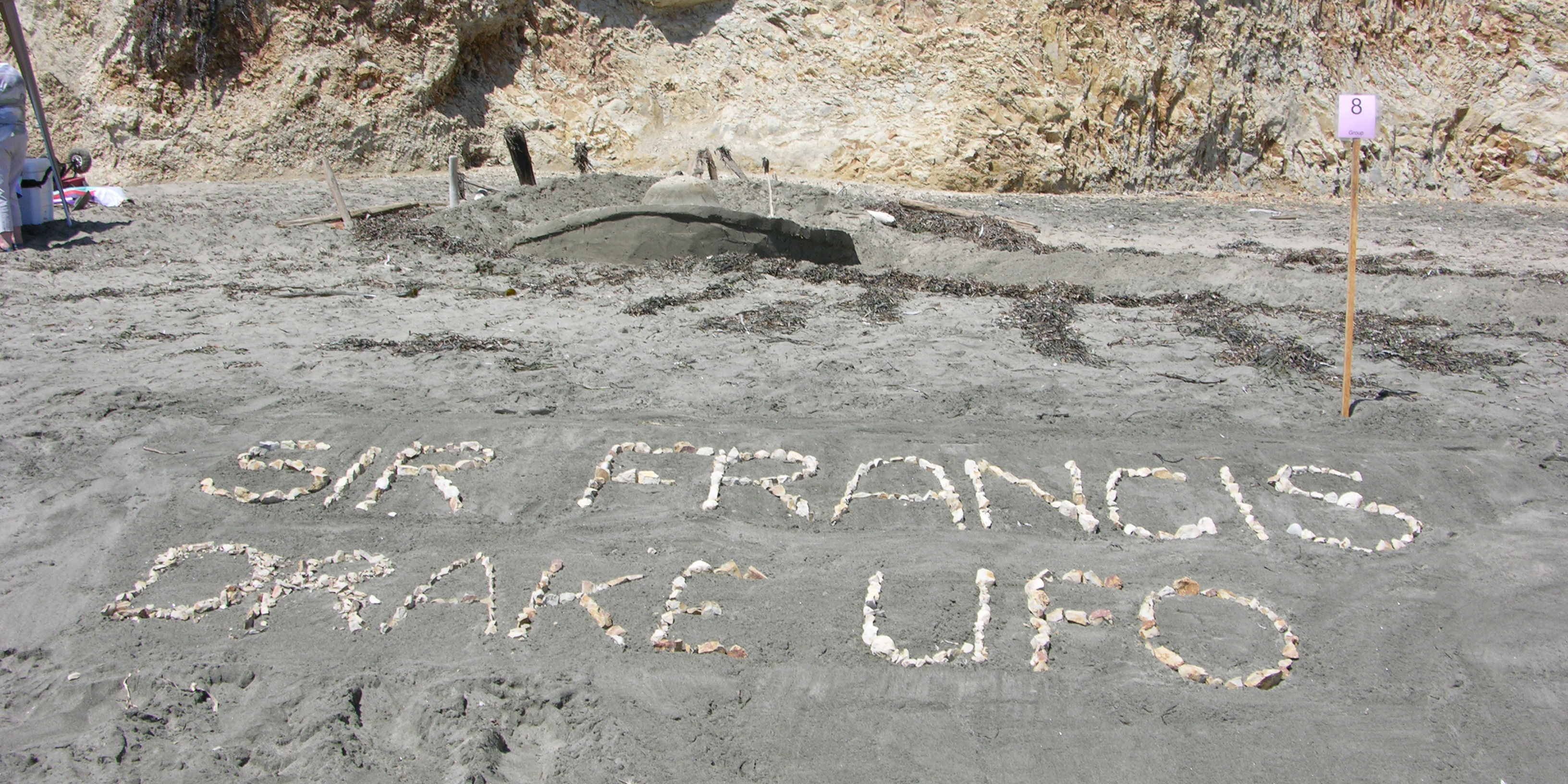 A large sand sculpture of a crashed flying saucer. In the foreground are beige rocks spelling out Sir Francis Drake UFO.