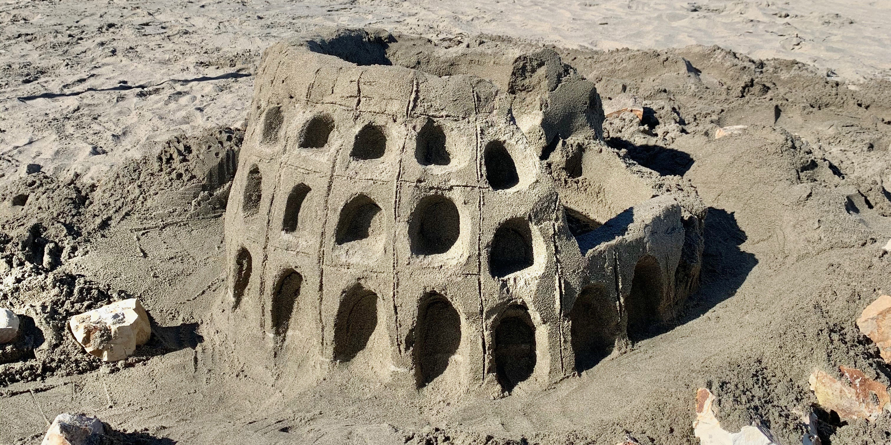 A small sand sculpture of the Coliseum in Rome, Italy. The sculpture is a bit more than a foot tall, is circular in shape with alcoves carved into the exterior walls, and part of the wall collapsed on the right.