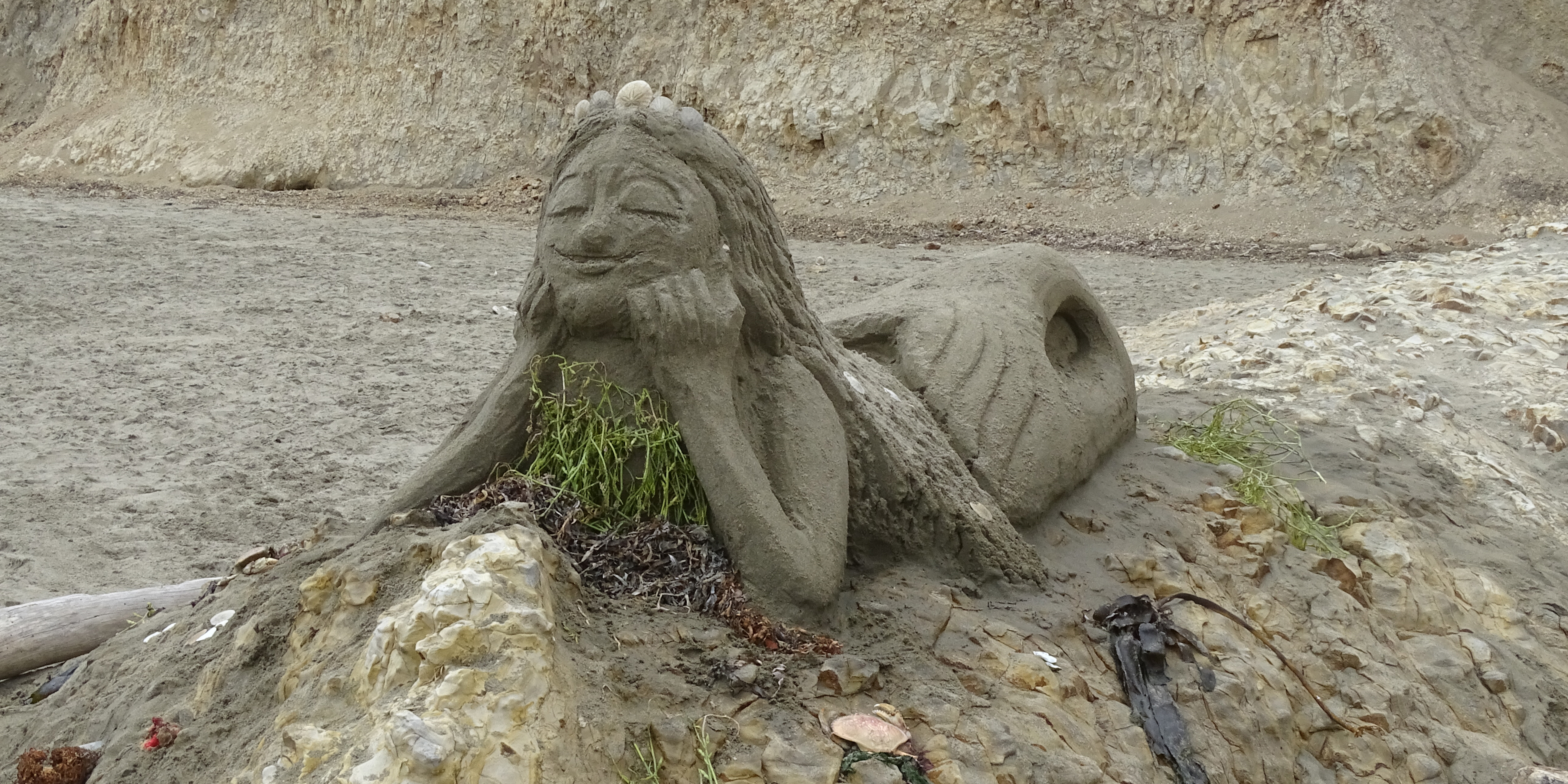 A sand sculpture of a mermaid lying on her belly on top of a rock outcrop with her elbows on the rock and head propped up in her hands.