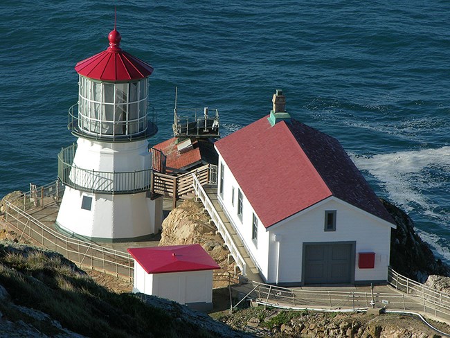 A white light tower with a red roof next to a few other white buildings on the edge of an ocean cliff.
