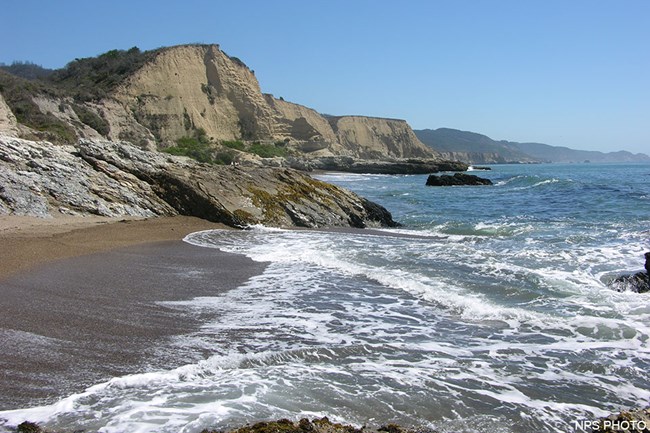 Rocky outcrops extend from tan-colored bluffs on the left into the ocean on the right.