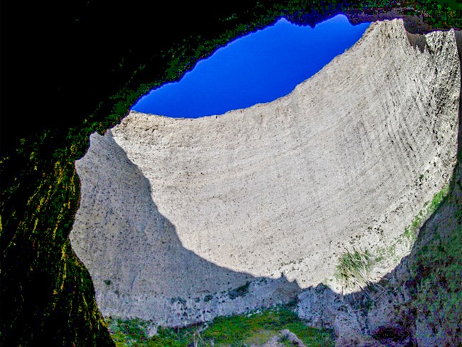 Looking up at blue sky visible through a large circular hole in the roof of a sea cave carved from beige sandstone.