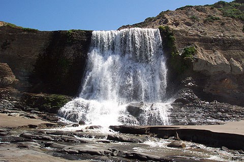Water falling over a 30-foot tall cliff top onto a sandy beach.
