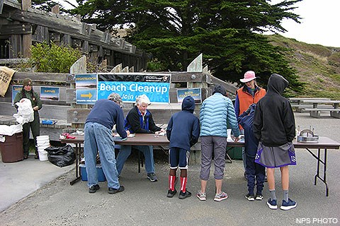 Five volunteers sign forms at a folding table while a ranger on the left prepares bags and buckets for the volunteers to use as they collect marine debris from the beach.