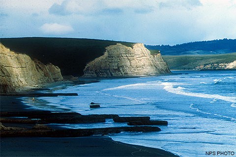 Sun-dappled, light-tan-colored cliffs on the left above a mostly sandy beach with waves washing ashore from the right.
