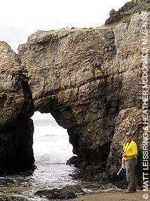 A man in a yellow jacket stands next to a small sea arch.