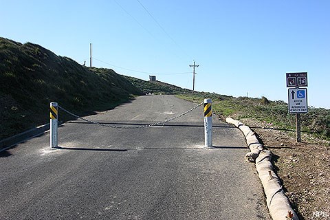 Two metal bollards connected by a heavy chain block a narrow road leading up a hill.