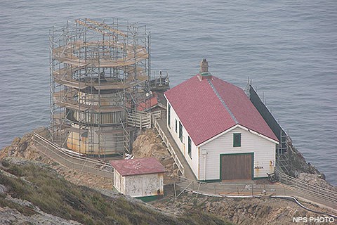 Three white-sided, red-roofed structures sit on a rocky headland above the Pacific Ocean. Scaffolding surrounds the lighthouse tower and has been erected on the right side of the larger building on the right.