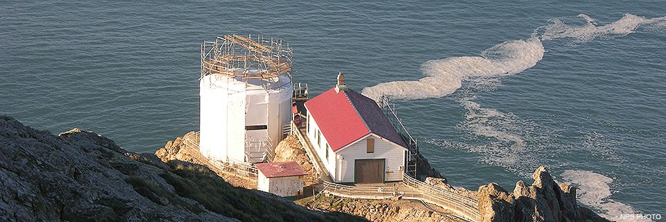 Three structures—a tower, a large rectangular building, and a small square structure—sit on a rocky headland above the Pacific Ocean. The roof and rafters of the lighthouse tower have been removed and scaffolding and white fabric surrounds the tower.