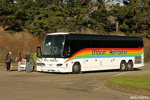 Five visitors boarding a coach-sized bus that is decorated with a rainbow-colored horizontal stripe.