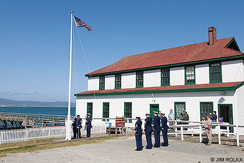 U.S. Coast Guard personnel and park visitors standing at attention and saluting as the U.S. flag is raised up a flagpole at the Historic Lifeboat Station at Chimney Rock.