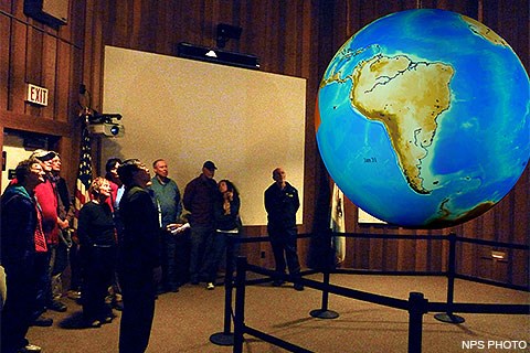 A park ranger and nine visitors looking at an image of Earth projected on a sphere which is hanging in the middle of an auditorium.