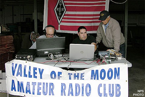 Three men sitting at a folding table on which there are laptops and radio equipment. A white banner with blue letters identify them as members of the Valley of the Moon Amateur Radio Club is hanging from the front of the table.