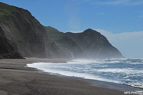Pacific Ocean waves wash ashore onto a sandy beach from the right. Bluffs rise from the beach on the left. A tall headland rises in the center.