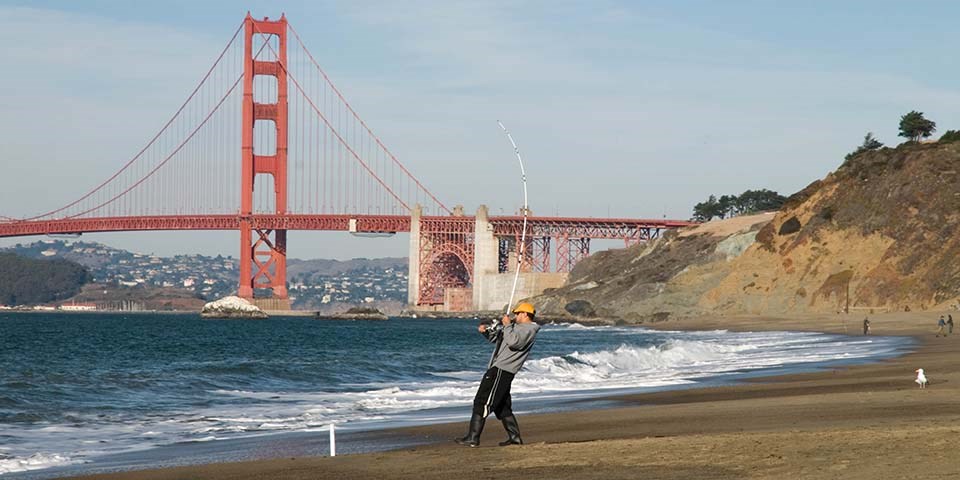 fisherman reels in his line on baker beach with bridge in the background