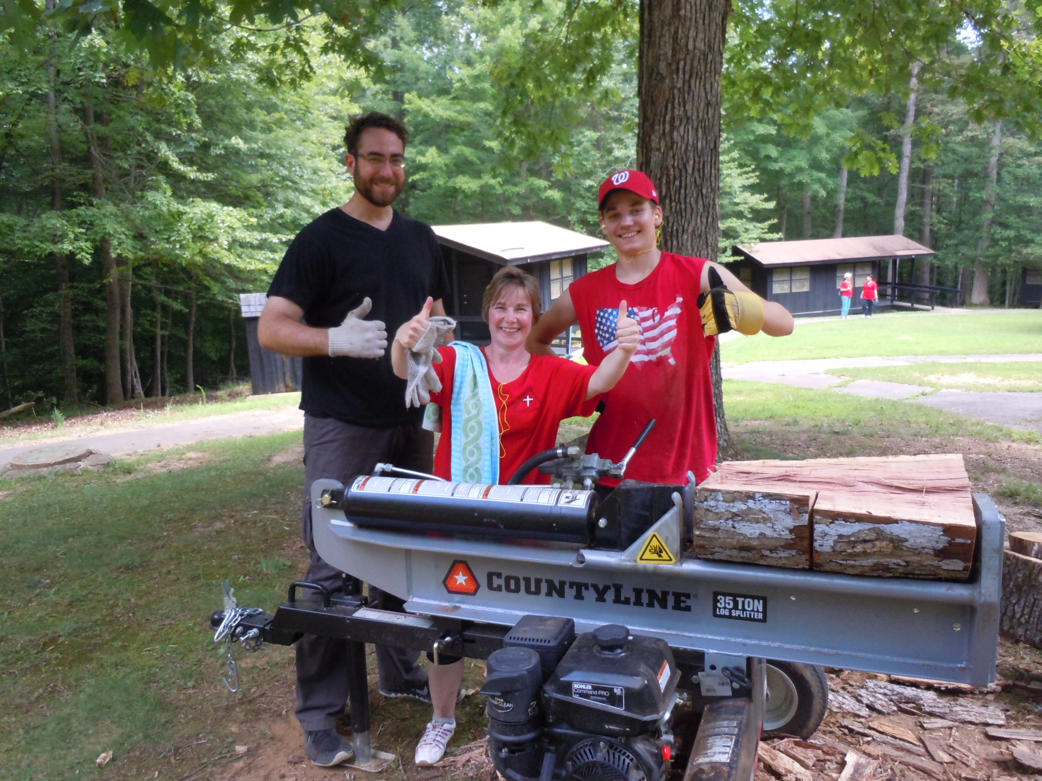 two adults and a youth use a log splitter to prepare firewood for campers