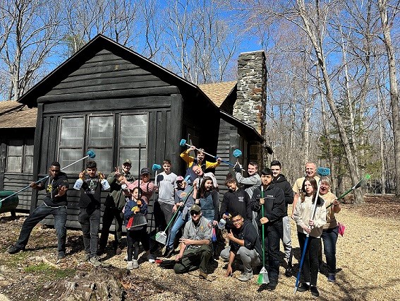 Group of people stand with brooms, dusters, and spray bottles in front of a wooden cabin in the forest