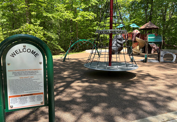 Children's playground structures near a welcome sign shaded by a green forest