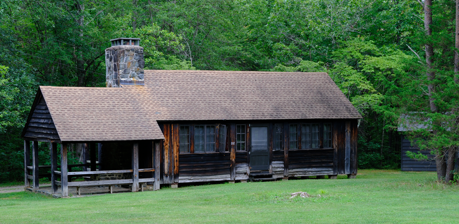 Wooden cabin building with a porch and a stone fireplace at the edge of a green forest