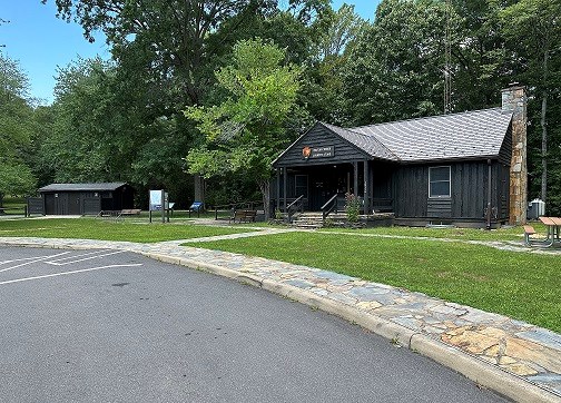 Wooden cabin like building at the edge of a green forest with a stone porch, stone chimney and stone pathways leading to it from a paved road