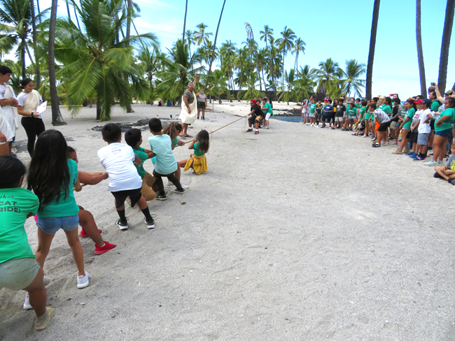 School children play huki huki (tug of war) upon the sandy shores of the Royal Grounds. A referee in a white kihei signals the start of the game.