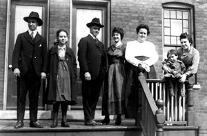 Seven Pullman residents on Champain Avenue stand on the porch of a brick rowhouse looking towards the camera. Two of them are children.