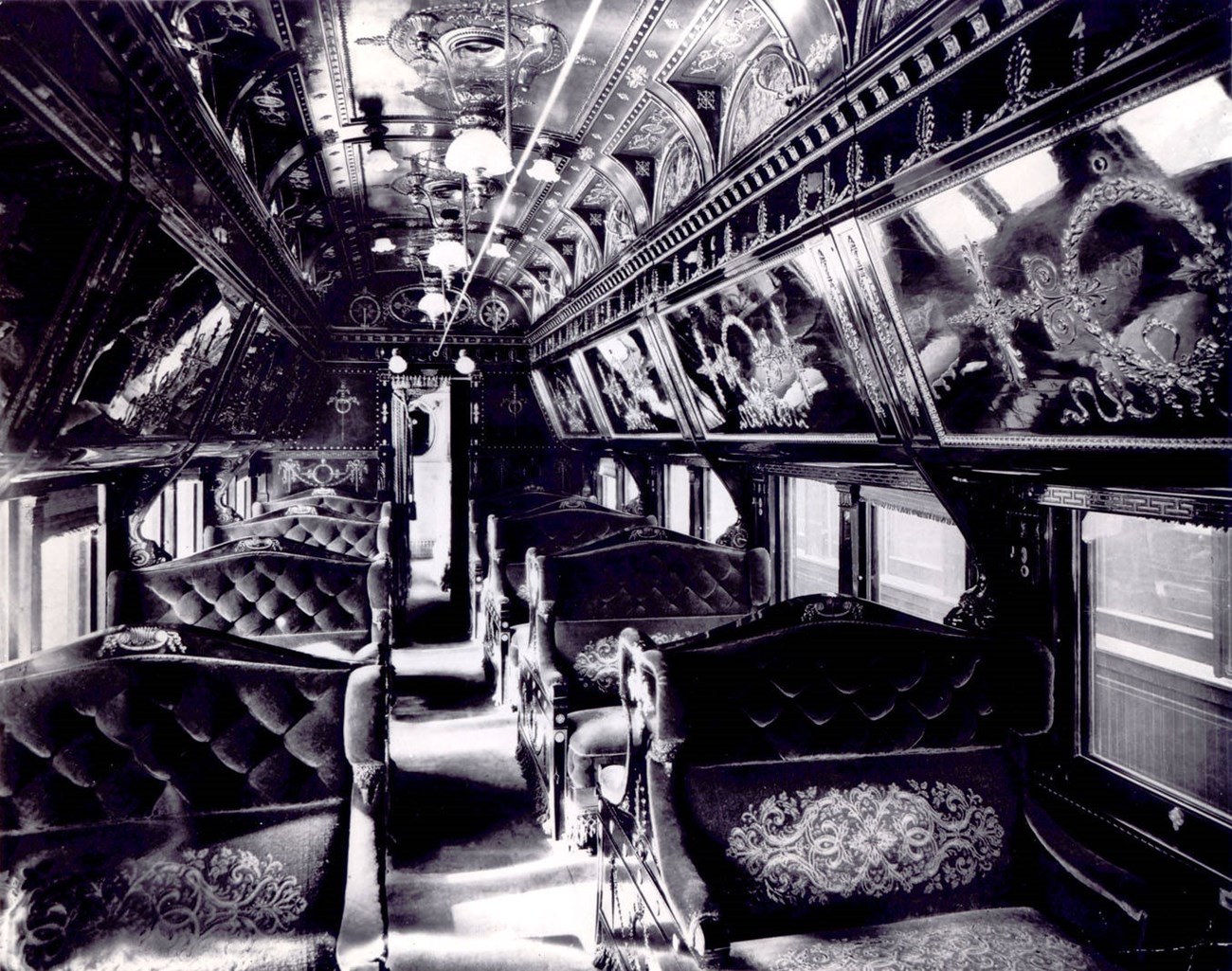 Interior of a train car with plush seating and carpeting and decorative luggage overheads, black and white photo.