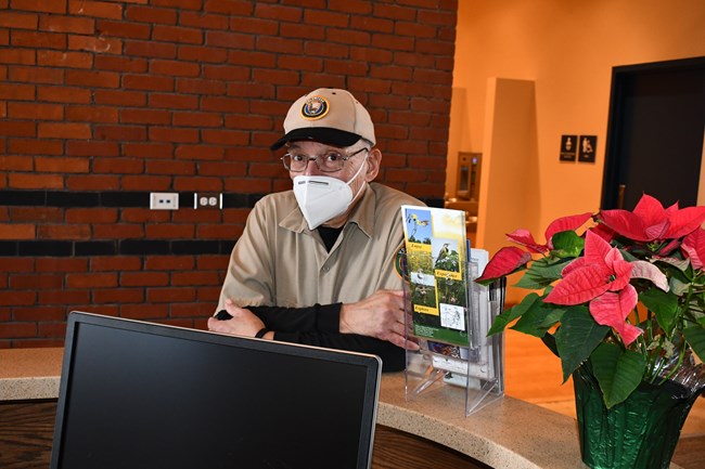 A man wears a mask and leans against a greet counter in a NPS volunteer uniform.