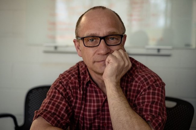 A man in a red polo rest his chin on his hand and smiles at the camera.