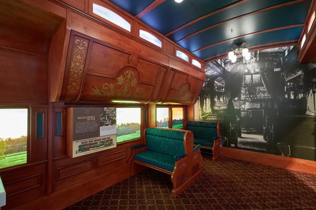 The inside of a replica train car with decorated wooden overhead bunk storage and cushioned green seats.