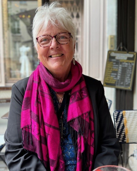 A smiling woman with white hair wearing pink tortoiseshell glasses, a pink scarf, and black jacket.