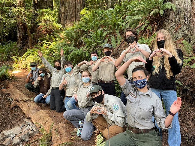 A crew of young adults and two park employees gather on a hiking trail. Redwood trees are behind them.