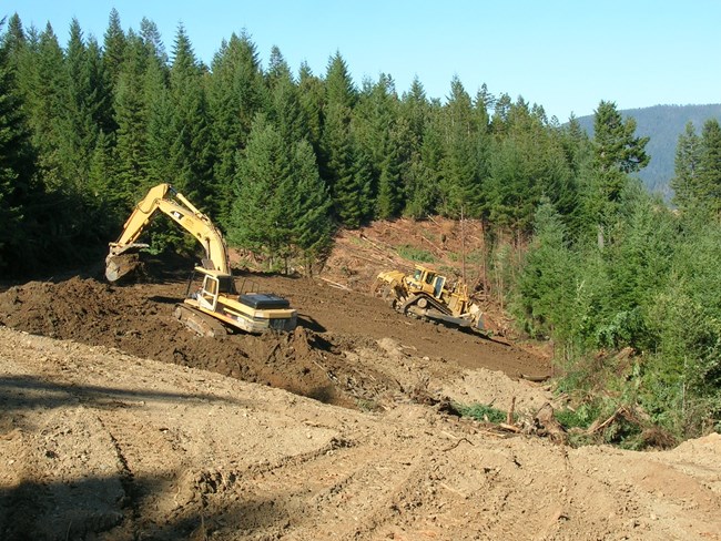 Heavy machinery removes old logging roads. Forests are in the background.