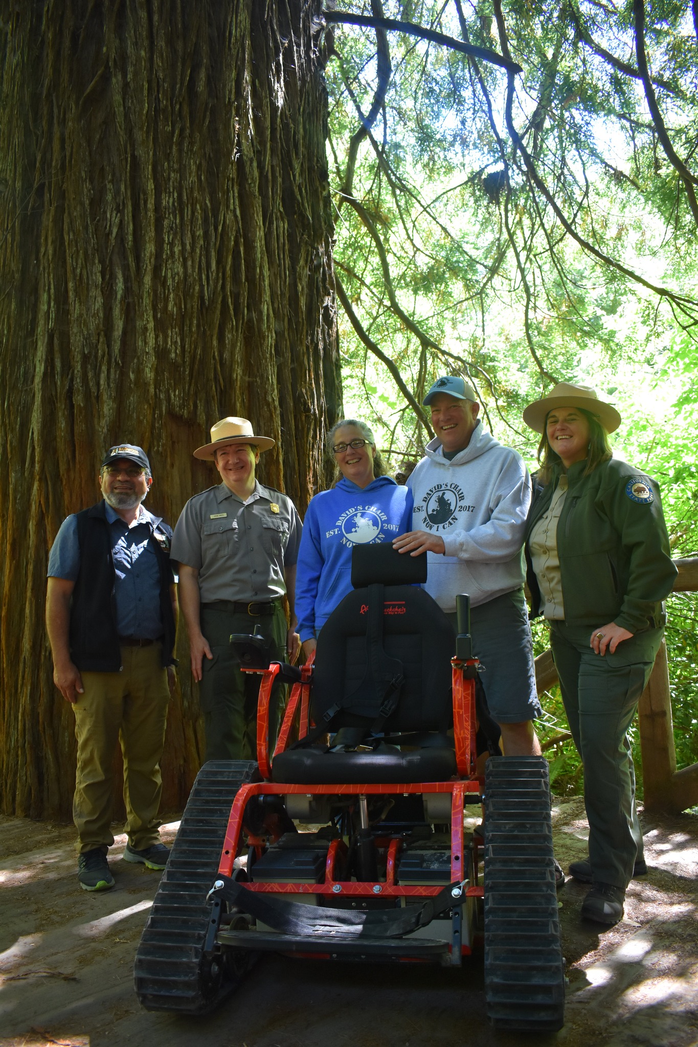Park staff stand with a red track chair on a trail in the redwoods