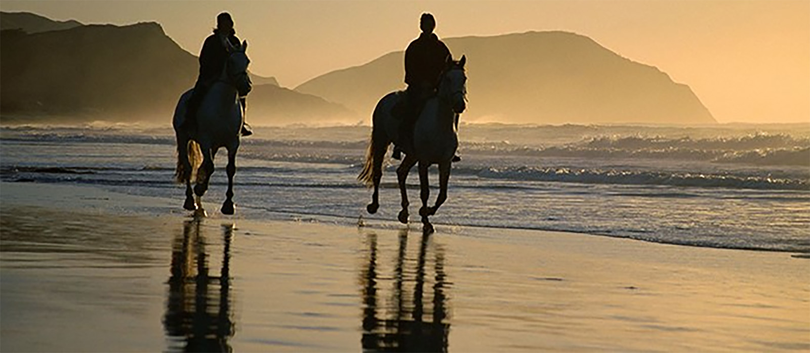 Two people riding horses on the beach at sunset.