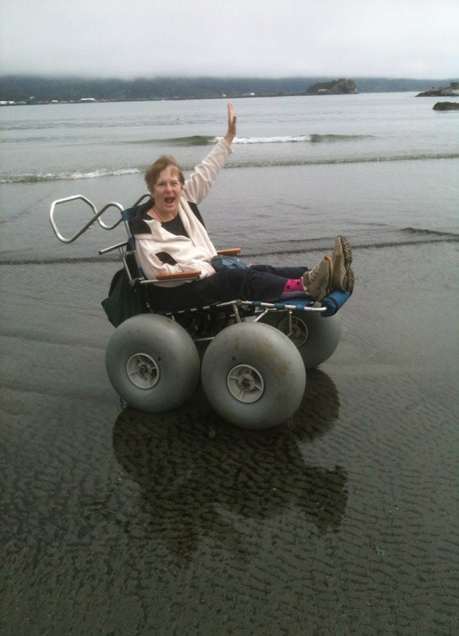 A woman in a beach wheelchair smiles and waves