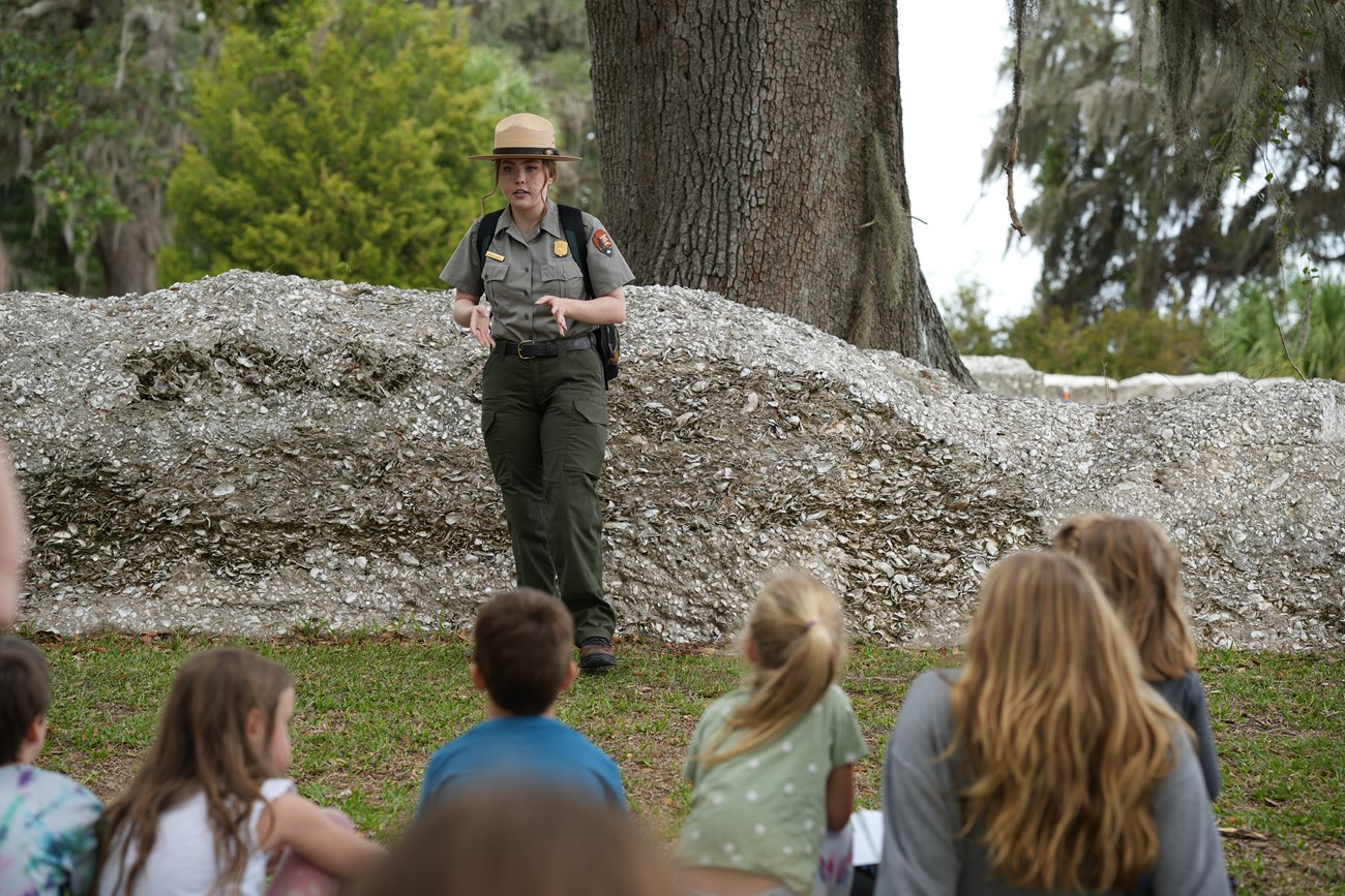 A ranger stands in front of ruins of a fort talking to a group of kids sitting on the group.